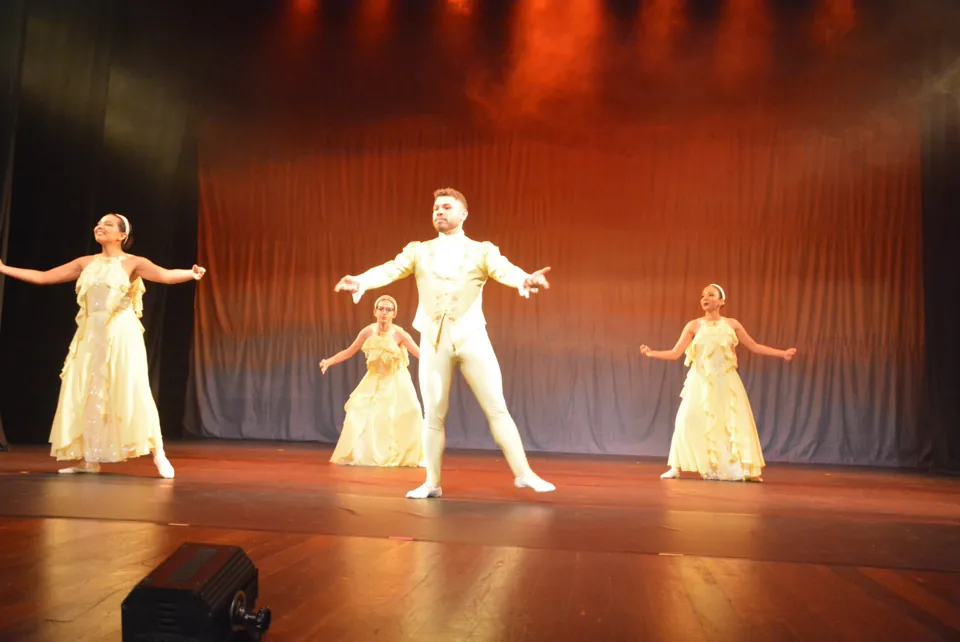 Bailarino de figurino claro ao centro do palco, com três bailarinas de vestidos amarelos, em coreografia de ballet adulto.