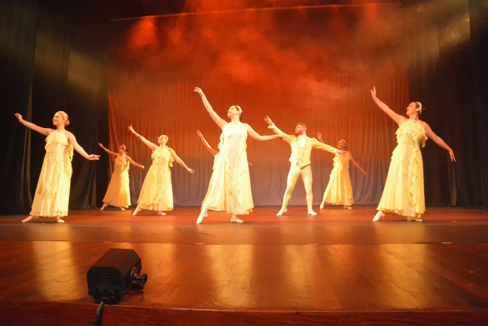 Turma de ballet adulto com vestidos amarelo-claros e um bailarino dança com os braços abertos em palco iluminado em tons de laranja.