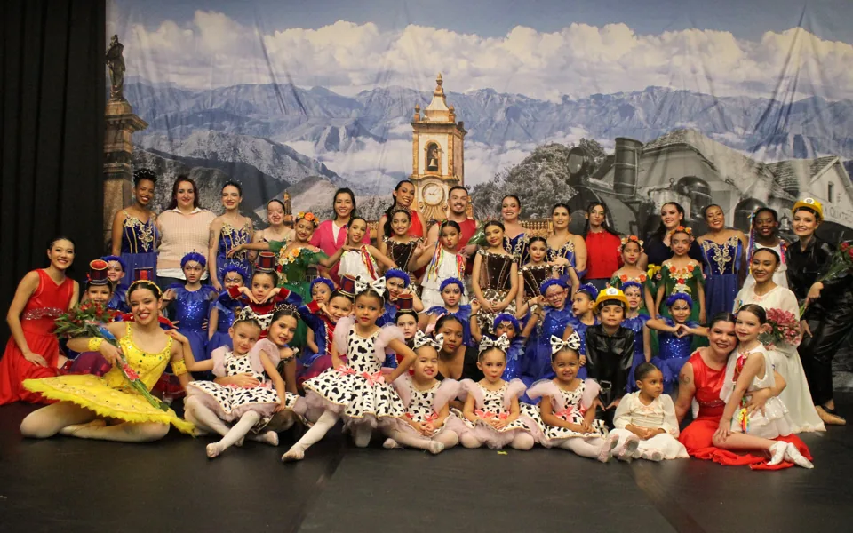 Elenco do Democratie reunido no palco do Trem di Dança, em 2025: alunas e alunos de todas as idades, de bebês a adultos, com figurinos coloridos diante de um cenário de montanhas, igreja e locomotiva.