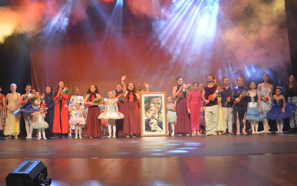 Elenco completo do Democratie agradece ao público no palco, sob luzes coloridas e fumaça, com flores nas mãos e um quadro em homenagem ao centro.