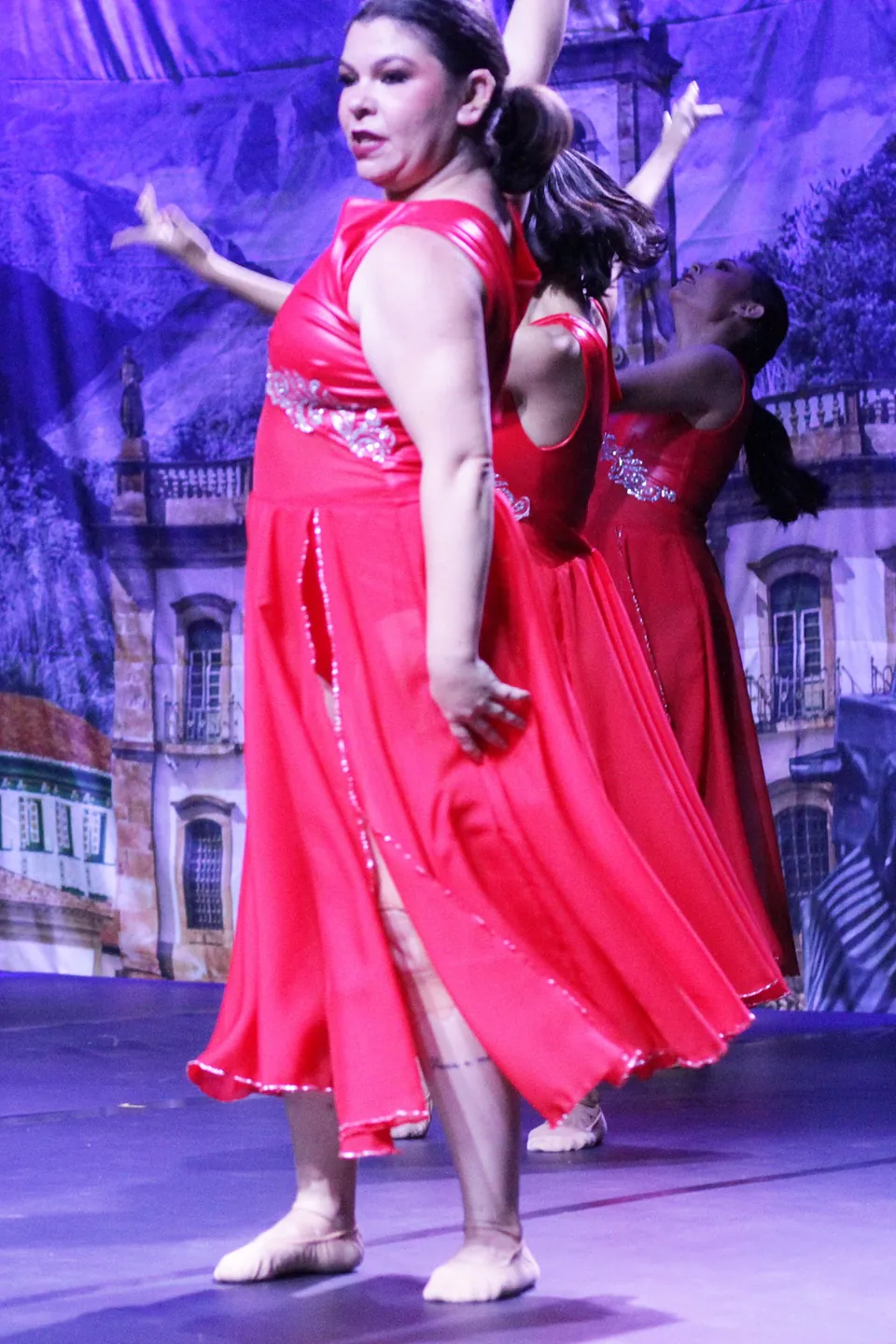 Bailarina de vestido vermelho gira em cena de jazz, com outras bailarinas ao fundo, no espetáculo Trem di Dança, em 2025.