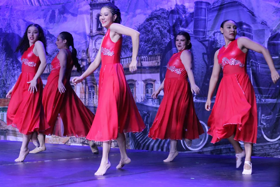Cinco bailarinas de vestidos vermelhos dançam jazz em sincronia diante do cenário do espetáculo Trem di Dança, em 2025.