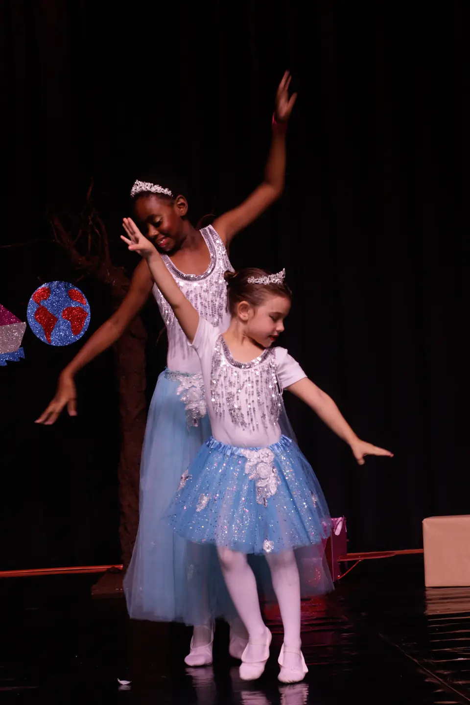Duas bailarinas com tutus azuis e tiaras, uma maior atrás da menor, abrem os braços em sincronia.