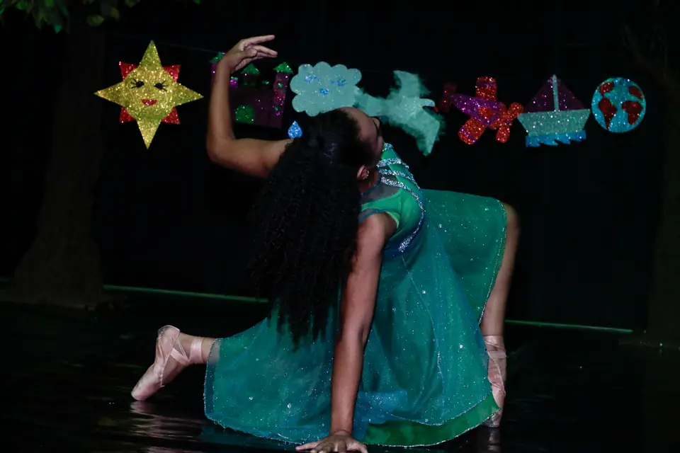 Bailarina de vestido verde e sapatilhas de ponta se curva em cena com sol, nuvens e barquinhos de papel ao fundo.