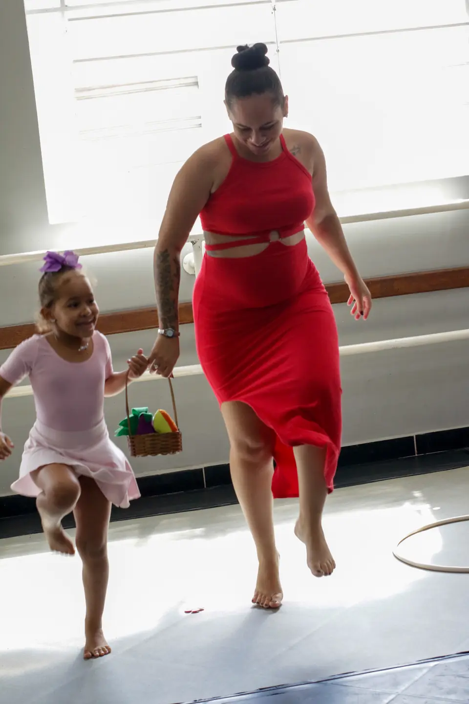 Na sala do Studio, uma menina de collant rosa e uma mulher de vestido vermelho dançam juntas sobre o linóleo, com barras de ballet ao fundo.