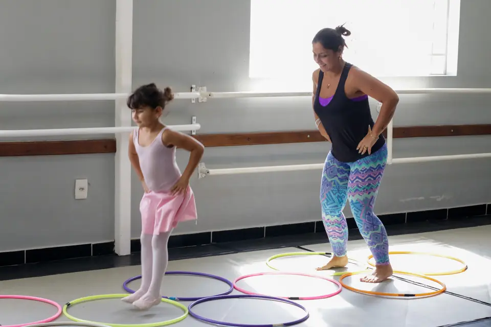 Sala de dança do Democratie com janelas amplas e barras: menina de collant rosa e mulher pulam entre bambolês coloridos no chão.