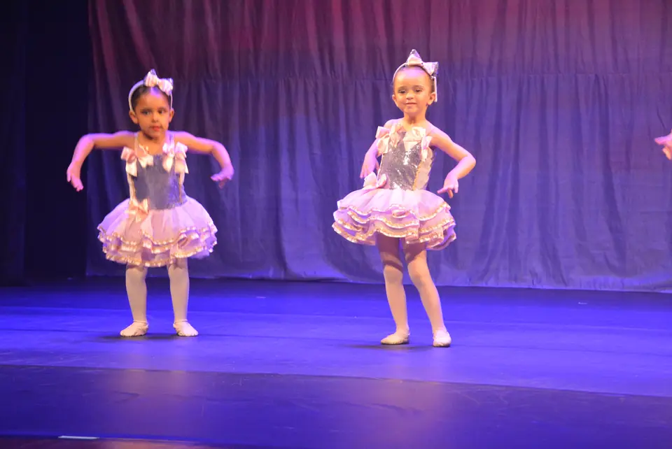 Duas pequenas bailarinas com saias de babados lilás e laços na cabeça dançam no palco iluminado de azul.