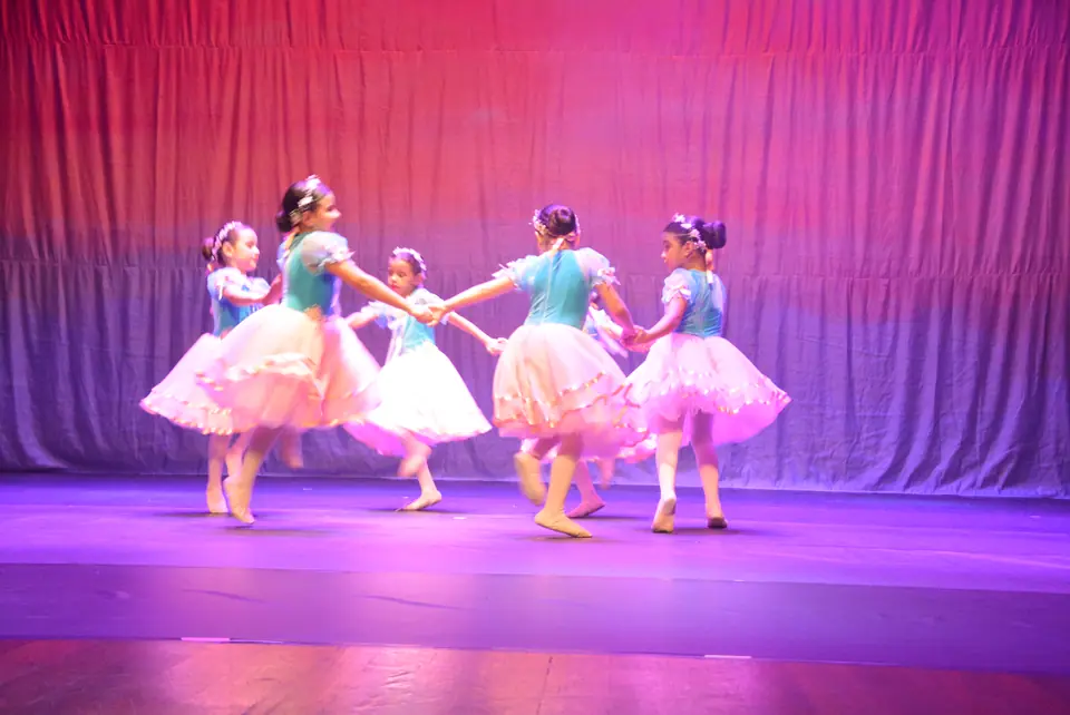 Crianças com tutus brancos e corpetes verdes dão as mãos em roda no palco iluminado de rosa e lilás.