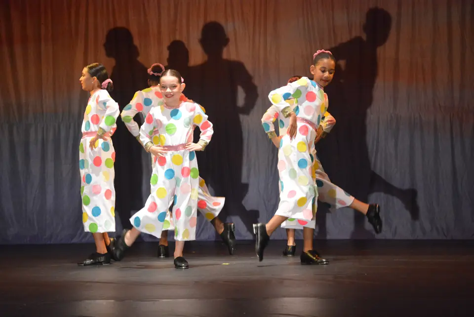 Jovens bailarinas com macacões brancos de bolinhas coloridas dançam em linha, com sombras projetadas na cortina.