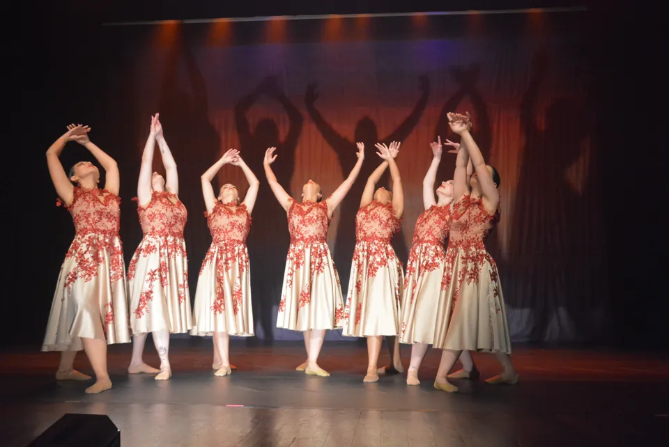 Sete bailarinas com vestidos claros bordados de vermelho erguem os braços em sincronia no palco do espetáculo Travessia, em 2026.