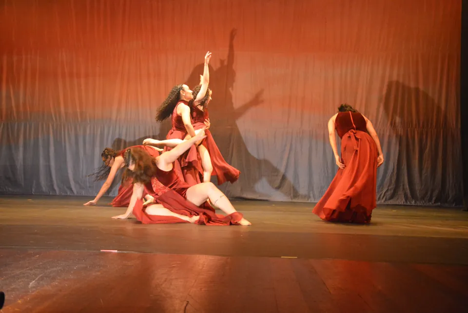 Bailarinas de vestidos vinho em cena de dança contemporânea, entre movimentos no chão e braços estendidos, no espetáculo Travessia, em 2026.