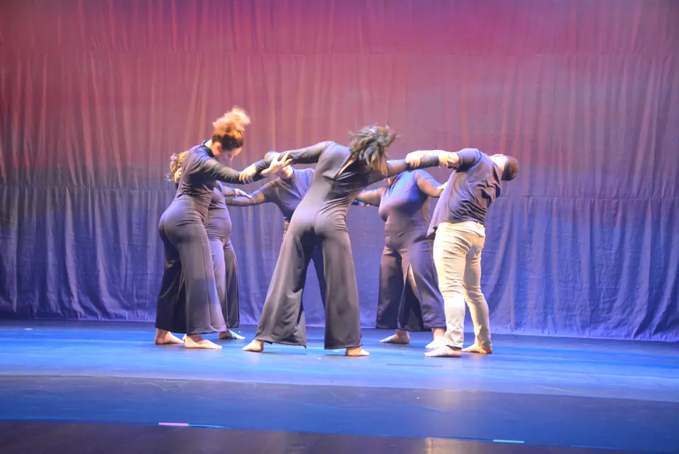 Grupo de bailarinos com macacões escuros, de braços entrelaçados, em cena de dança contemporânea sob luz azul e rosa.