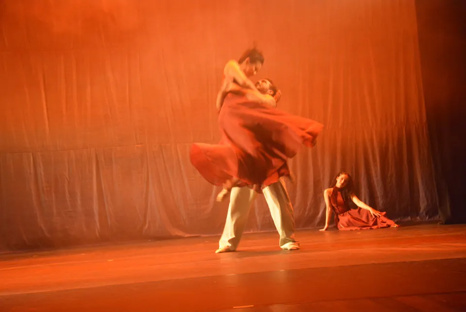 Bailarino ergue a parceira, de vestido vermelho esvoaçante, sob luz alaranjada no espetáculo Travessia, em 2026.