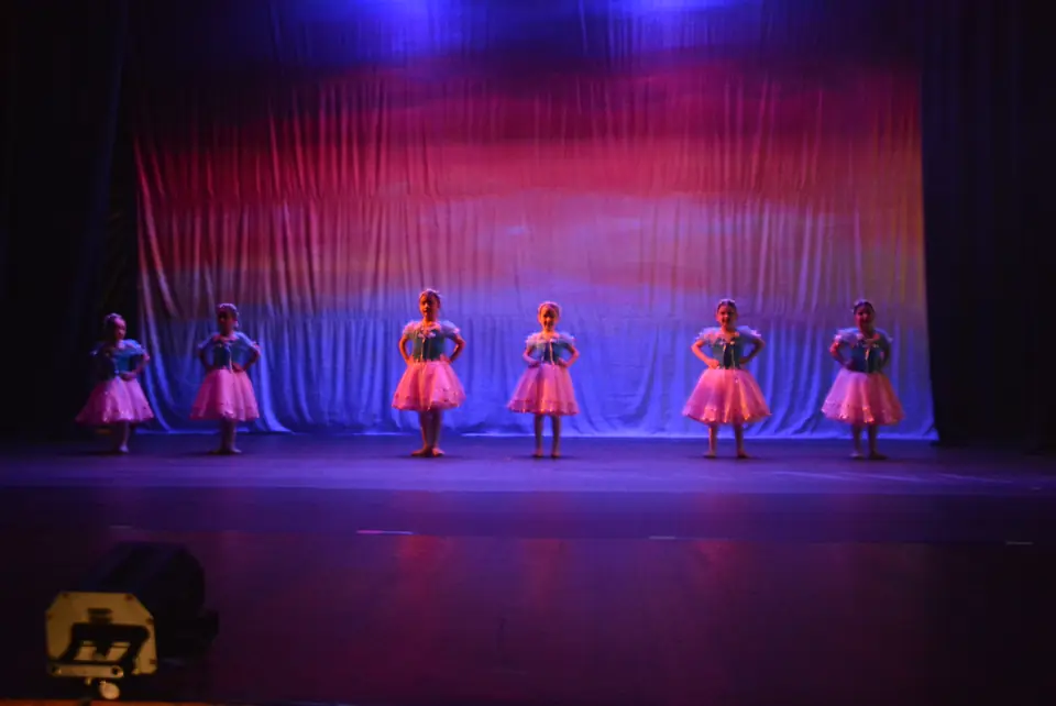 Fileira de pequenas bailarinas com saias cor-de-rosa no fundo do palco, sob iluminação azul e magenta.