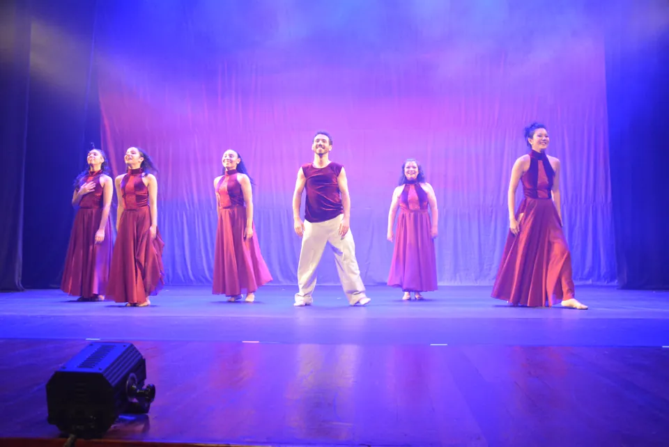 Elenco adulto alinhado no palco iluminado em azul e lilás, ao fim de uma coreografia do espetáculo Travessia, em 2026.