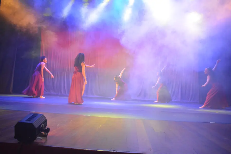 Bailarinas com vestidos vermelhos atravessam a névoa iluminada por refletores coloridos.
