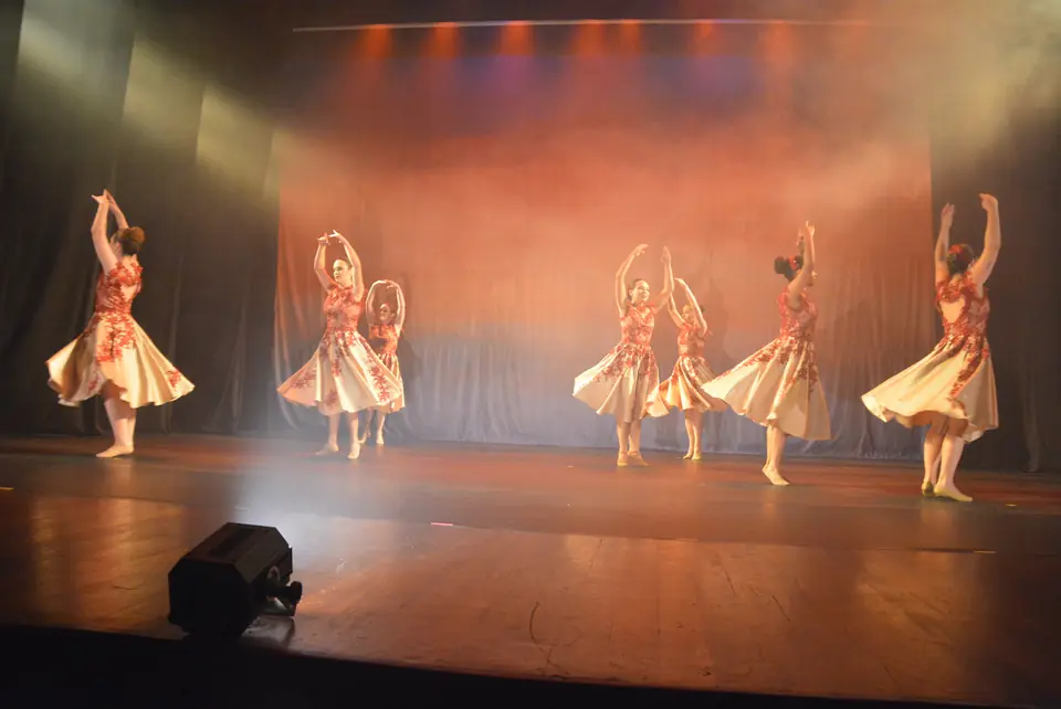 Palco do espetáculo Travessia, em 2026, visto de longe: bailarinas com saias claras e corpetes vermelhos dançam entre névoa e luz alaranjada.