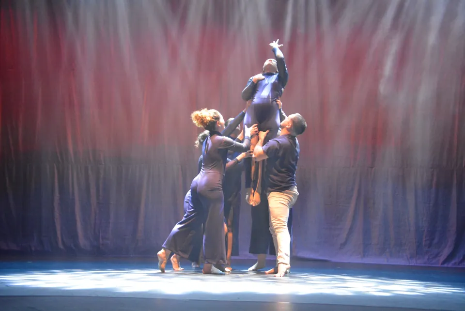 Grupo de bailarinos de azul-marinho sustenta uma bailarina no alto, que estende o braço para cima, no espetáculo Travessia, em 2026.