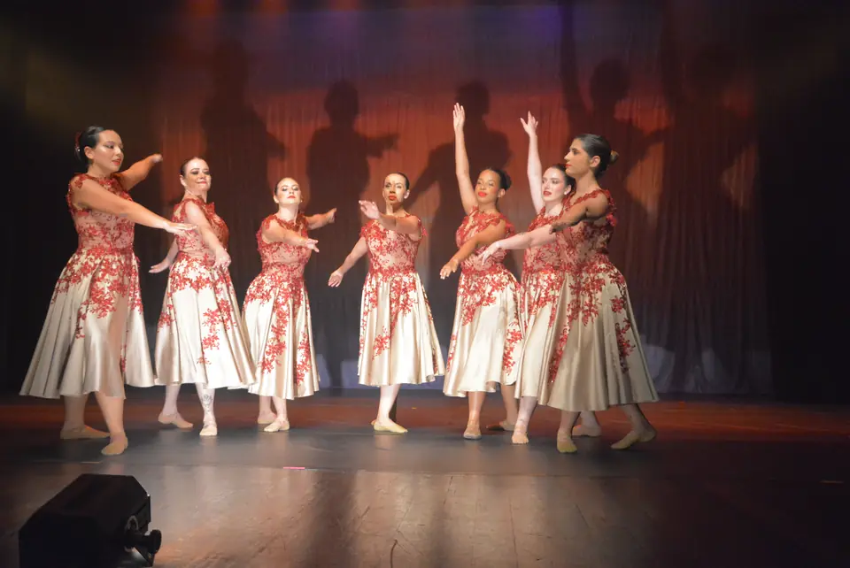 Bailarinas com saias brancas estampadas de flores vermelhas posam em conjunto, com braços elevados.