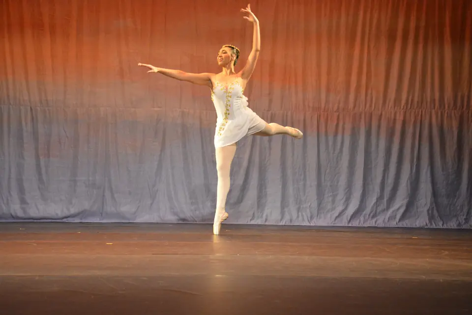 Bailarina solista de figurino branco executa um arabesque na meia-ponta, com os braços abertos.