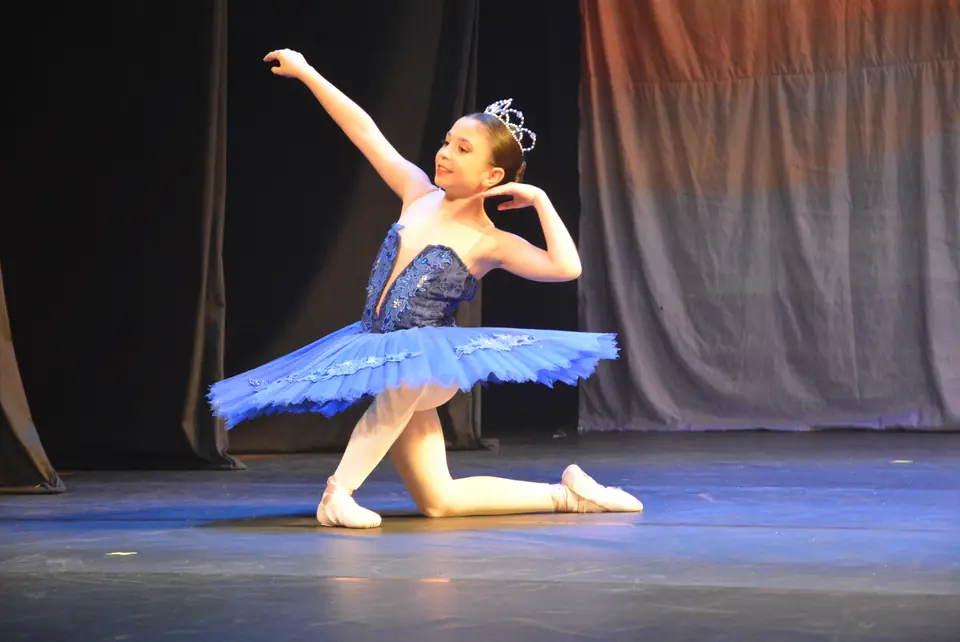 Jovem bailarina com tutu azul e tiara ajoelhada em pose de ballet clássico, com um braço erguido.