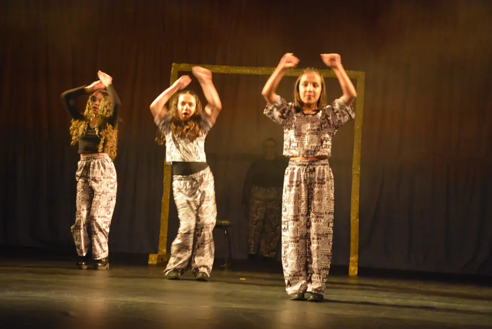 Três jovens com calças estampadas em preto e branco erguem os braços em coreografia de estilo urbano.