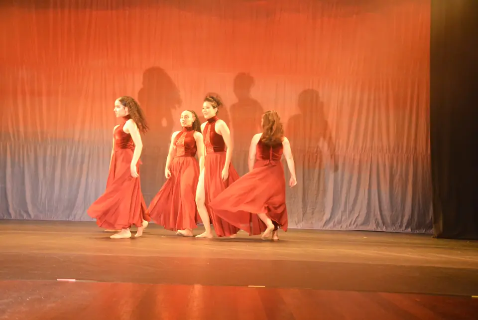 Quatro bailarinas com vestidos longos vermelhos em cena de Travessia, diante de cortina iluminada em tons de laranja.
