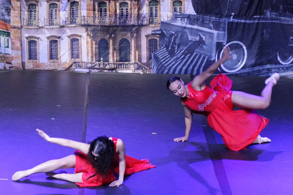 Duas bailarinas de vermelho em movimentos de chão diante de um casarão colonial pintado no cenário do Trem di Dança, em 2025.