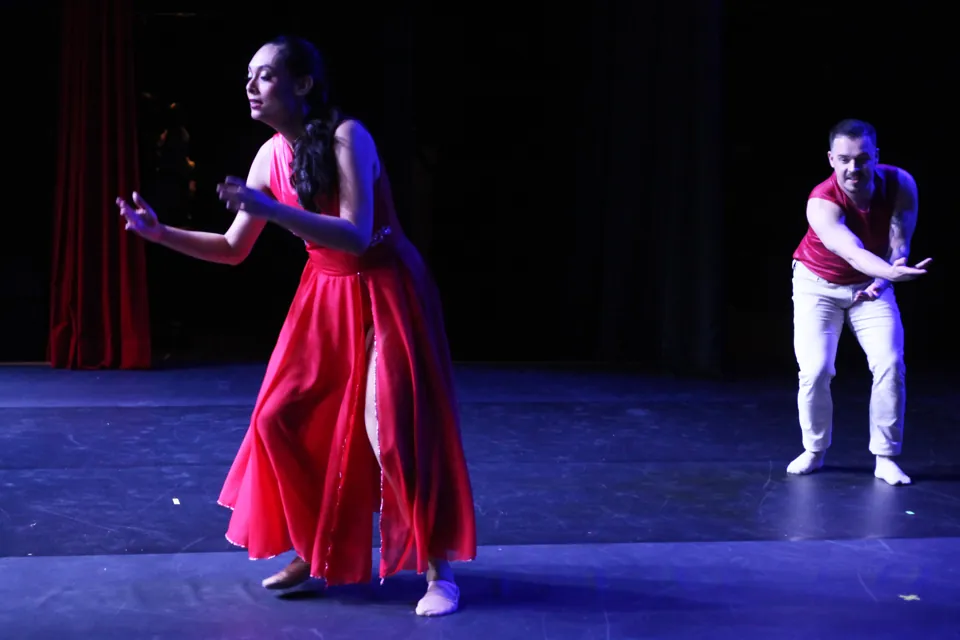 Bailarina de vestido vermelho e bailarino dançam frente a frente no palco escuro do espetáculo Trem di Dança, em 2025.