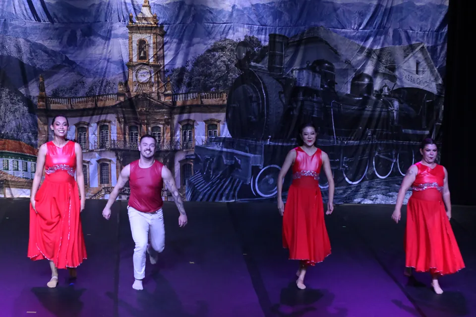 Bailarinas de vestidos vermelhos e um bailarino dançam diante de um cenário com igreja colonial e locomotiva, no espetáculo Trem di Dança, em 2025.