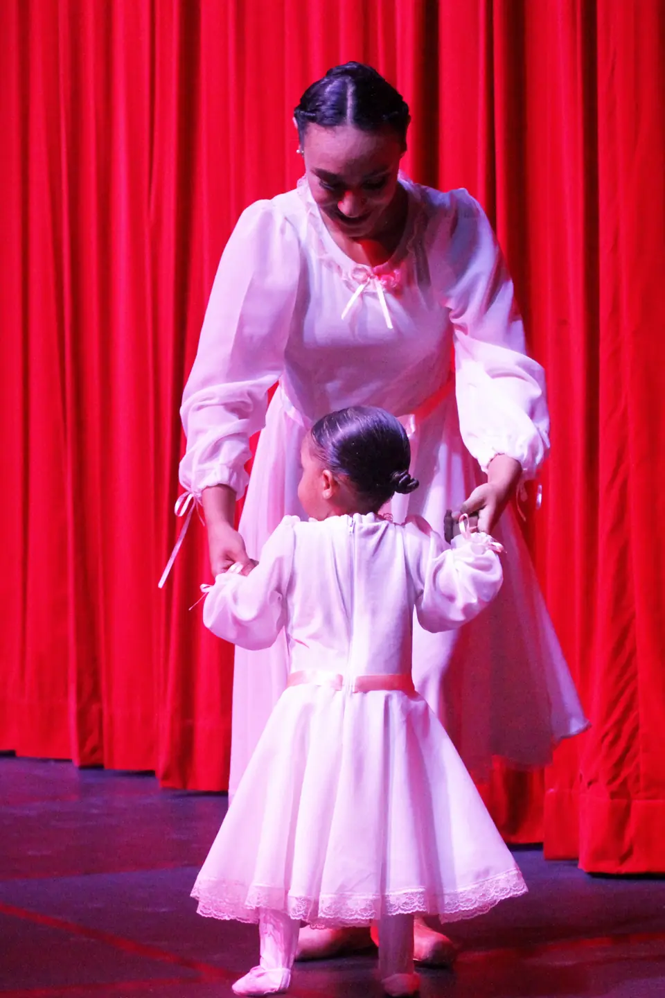 Mulher de vestido branco conduz pelas mãos uma criança pequena de vestido branco diante de uma cortina vermelha.