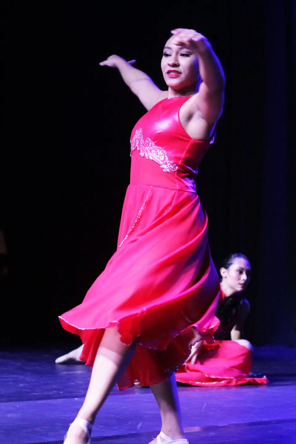 Bailarina de vestido vermelho gira em primeiro plano enquanto outra intérprete aguarda sentada ao fundo, no Trem di Dança.